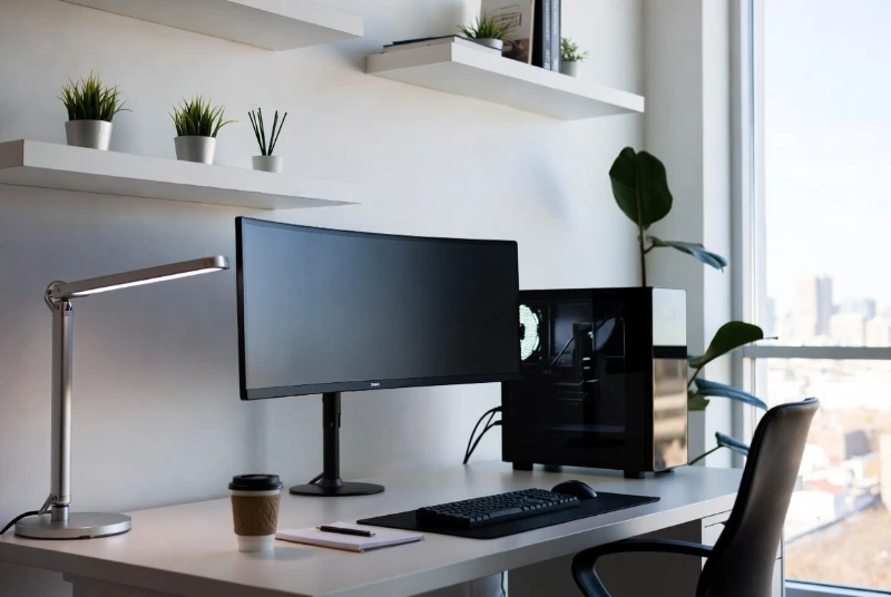 Clean professional desk with ultrawide curved monitor, desktop PC, LED lamp, and city view through large window